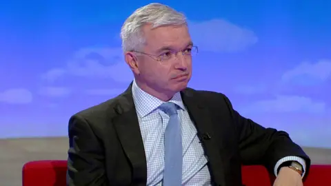 BBC Mark Menzies is interviewed by the BBC's Sunday Politics North West programme in 2018. He is wearing a blue tie and black suit and sits on a red sofa in front of a blue backdrop.