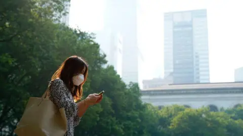 A woman walks through New York wearing a mask and looking at her phone while smog from wildfires hangs over buildings and skyscrapers in the city