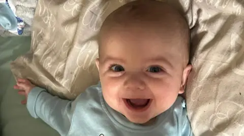 Family photo Jensen-Lee laughing in a close up shot. His mouth is open as he smiles. His head is resting on a beige pillow and green bed. 