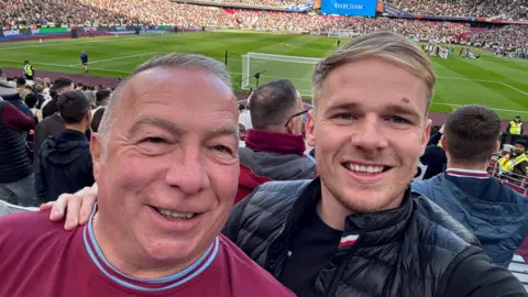 Kevin Gray (left) in a West Ham shirt smiles and looks at the camera. His son Jamie (right) in a black coat has his arm around him and is also smiling. There is a football pitch in the background.