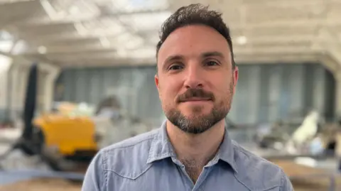 Emma Baugh/BBC Adrian Kerrison is standing in a large plane hangar which has an aircraft to the left. He has dark brown hair and a short beard and is wearing a pale blue denim shirt.