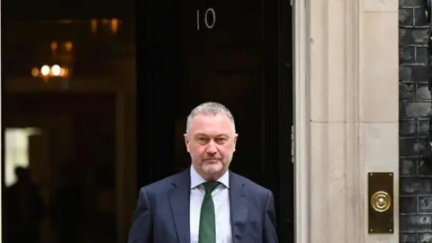 Getty Images Steve Reed is seen leaving 10 Downing Street. He is wearing a dark navy suit with a green tie while holding a red folder.