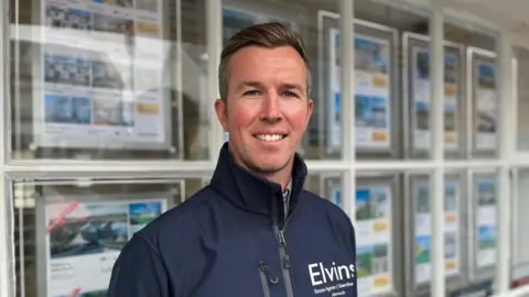 Rhys Elvins, looking at the camera, standing in front of an estate agents window and is wearing a blue top which has Elvins written on it 