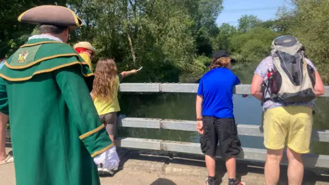 BBC Children drop sticks off the side of the bridge. A man in traditional clothing, like a town crier, looks on, commentating. 