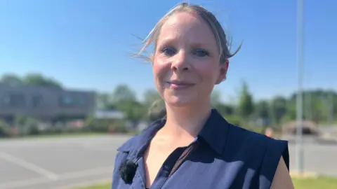 Emma Baugh/BBC Vikki Johnson smiles at the camera while standing outside in the playground. She wears a sleeveless navy shirt and has tied-back blonde hair.