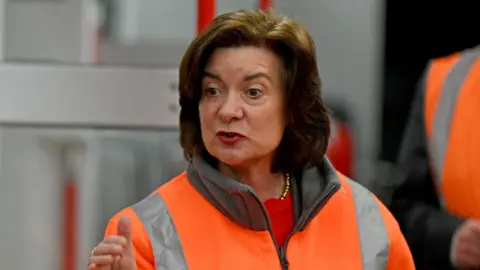 Eluned Morgan wearing a hi-viz jacket, guestering with one hand while she speaks. She is stood in front of a train which is out of focus.