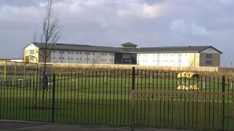 Chris Stafford/Geograph Exterior image of a HMP Peterborough wing, a white building with a dark roof, with a park in the foreground