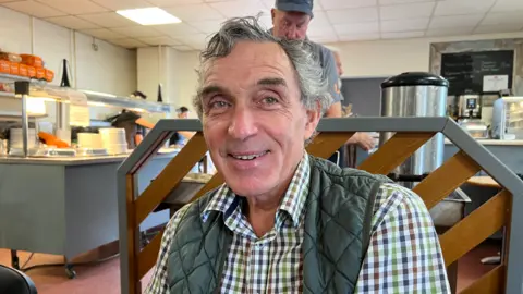 A man with wavy grey hair, a white checked shirt and green gilet sitting in a cafe with other men 
