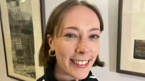  Clara Kerr. She has short brown hair and blue eyes. She is wearing gold hoop earrings and a black jacket. Pictures are behind her.