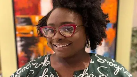 Seyi B Seyi is wearing a dark green blouse and smiling at the camera. She has big red glasses frames and neatly styled hair.