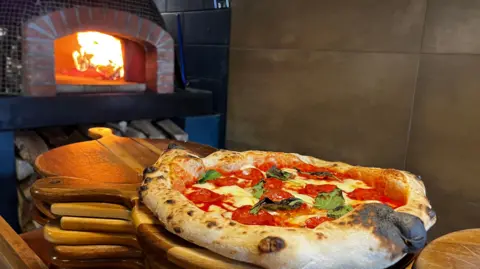   Una pizza fresca con salami y albahaca reposa sobre tablas de madera, justo delante de un horno de leña, con las llamas lamiendo el techo.