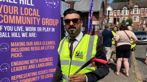 A man in a suit and sunglasses, with a hghi-vis vest, holds a litter picker while standing in front of a purple banner which has the words "YOUR LOCAL COMMUNITY GROUP" in large orange letters.