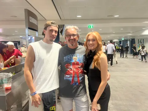 A picture of Paul and his daughter Alex and son Thomas smiling before the Bruno Mars concert. Paul is wearing Bruno Mars merch, Alex is wearing an all black outfit and Thomas is wearing a white tank top.