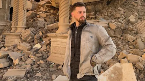 We Hate The Cold Harry Jackson is standing by ruins in Iraq. He is looking to the right of the frame and standing by a large pile of rubble. He is wearing a light-coloured jacket, brown gilet and has short dark hair and a beard. 