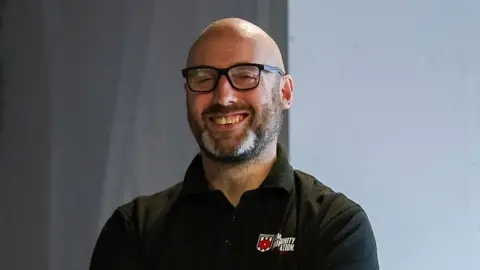David Airey Head and shoulders shot of Darren Jenkinson. He is standing in front of a grey curtain and has a bald head, dark-framed glasses and a salt and pepper short beard. He is smiling and wearing a black polo shirt