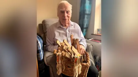 Brunelcare John is sitting in an armchair with a basket full of wooden monkeys in his lap. He is smiling at the camera.