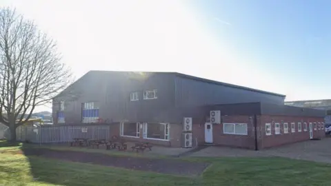 A factory building with bricks at the bottom and a dark grey paneled roof. In front are picnic benches and a fenced area.