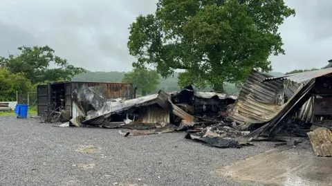 A farm shop building that has been destroyed in a fire. The rubble is now piled up on the floor. A large tree is behind the pile of debris.