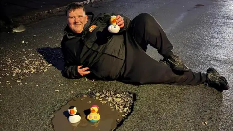 A man wearing a black jacket and trousers lies in the middle of the road at night, holding a white rubber duck. Two rubber ducks can be seen in a water-filled pothole in front of him.