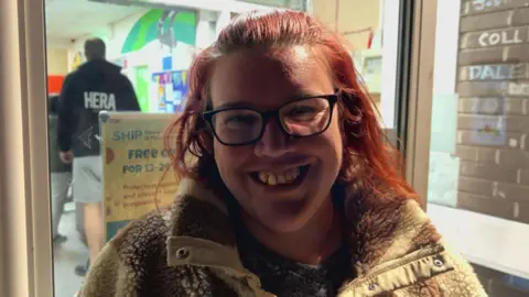 A woman smiling and wearing glasses. She has red shoulder length hair. She is wearing a fleece. She is stood in front of a glass door. 