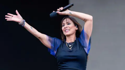 Natalie Imbruglia waves her hands over her head, with one hand clutching her microphone, as she performs on stage. She has a few tattoos, including one of what appears to be a seagull feather, on her arms, and is smiling. She is wearing a black dress with electric blue, puffy, mesh short sleeves. 