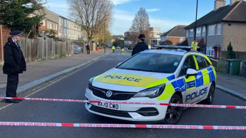 A police car and police tape at the location of the shooting. A female officer stands near the car with her back to the camera and a male officer stands to one side. More police can be seen further down the road.