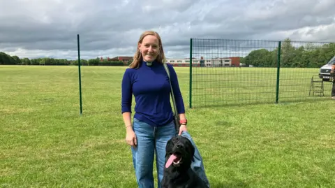 JOANNA TAYLOR/BBC The Reverend Allison Williams is smiling at the camera. She is wearing a navy jumper with a dog collar and blue jeans. Next to her is a black labrador with its tongue lolling out. Behind them is a partially built green metal fence over grass. 
