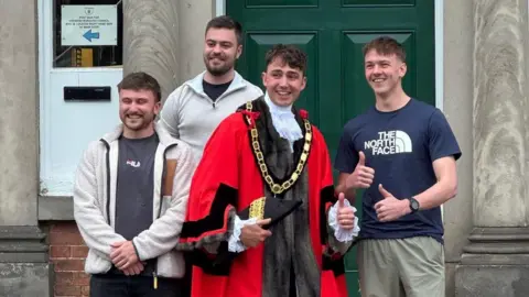 supplied Harry is smiling with his thumbs up. He is wearing red mayoral robes with black cuffs and a frilly white collar. He has a gold chain around his name. Stood beside him are three of his friends all dressed casually and also smiling at the camera. behind them in a green door.