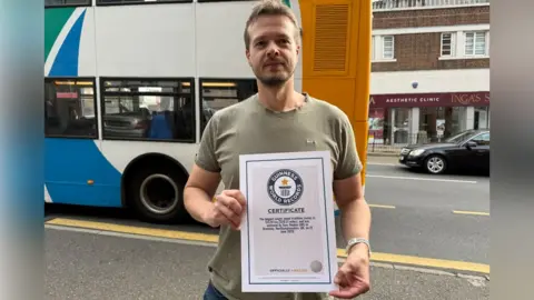 Robert Constantin/BBC A man in a green T-shirt holding a world record certificate. He is in front of a road where a bus and car is going past.