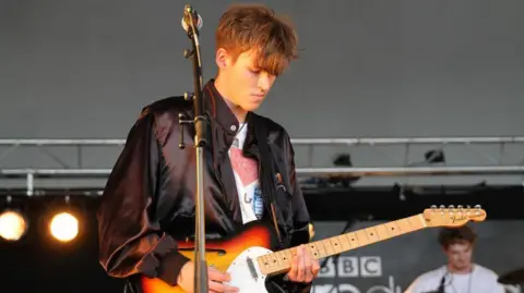 A man with short brown hair and a long fringe and wearing a white T-shirt and a black satin jacket is looking down while playing a Fender guitar on a stage. A man in a white T-shirt can be seen blurred out in the background, along with the top of a BBC Introducing Stage sign.
