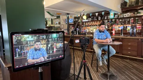 Kieran Doherty A Coventry City fan called Tommy wearng a Coventry City top, jeans and trainers, sits on a high stool with his hands crossed on the table in a pub. A camera on a tripod is to his left, and a microphone. A bar with drinks is behind him. A screen showing his face is further to the left infront of a wall.