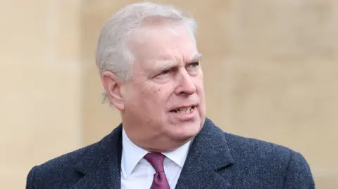 Prince Andrew stands outside in a grey suit with a maroon tie.