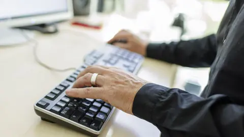 A person with a black long-sleeved shirt on is typing on a black keyboard connected to a computer screen. The person has a thick silver ring on their ring finger, and is moving a mouse with their right hand