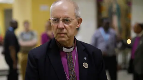 PA Media Archbishop of Canterbury Justin Welby looks on as he speaks with the press after a visit to the grave of Saint Oscar Arnulfo Romero, during a visit to El Salvador. Welby is wearing a black blazer with a bright purple shirt. A silver cross dangles from his neck against his chest.
