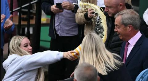 Woman throwing milkshake in Nigel Farage's face. He is wearing a black blazer with a pink shirt an tie