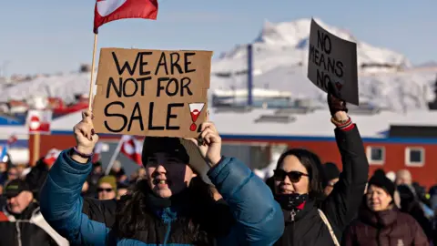 A protester holds a placard reading 'We are not for sale', with other protesters behind them against a backdrop of snowy mountains and the Greenland flag.