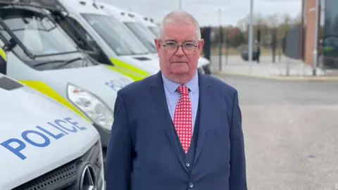 Laura Foster/BBC John Tizard staring into the camera while he is standing in front of police vans outside a police headquarters. He has short white hair and rounded glasses and is wearing a navy three-piece suit with a light blue shirt and red tie with silver spots. 