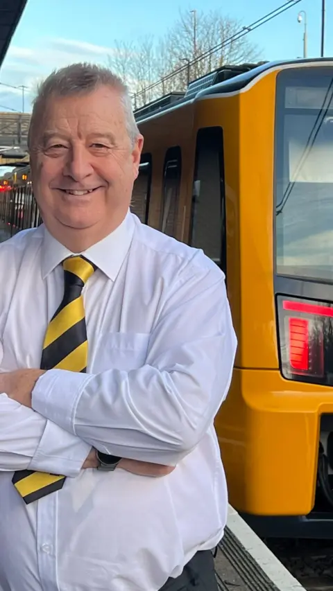 Metro driver Phil Taylor smilig with his arms folded. He is wearing a white shirt and a yellow and balck tie. He is standing on a Metro platform in front of a yellow and black Metro