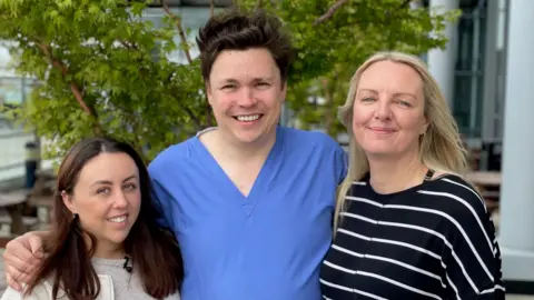OLIVIA RICHWALD/BBC A man in scrubs stands in the middle of two women with his arms around their shoulders. The woman on the left has long brown hair, the woman on the right has long blond hair. All three are smiling.