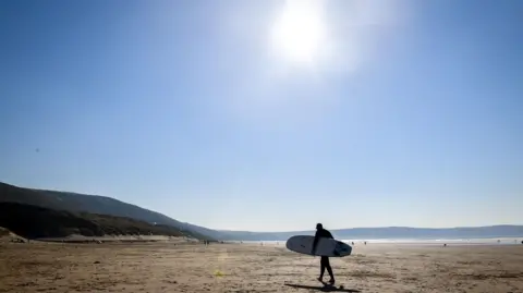 A surfer holding a longboard under his arm walks across a large empty beach. The sky is clear and blue and the sun is shining. The sea is glittering in the distance.