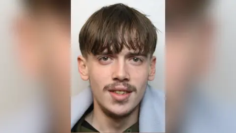 Wiltshire Police Leo Morris looking at the camera in a police mugshot. He has a long fringe that looks greasy and a straggly moustache. He has yellow teeth.