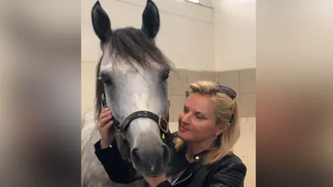 Contributed Ms Kelleway is pictured with a grey horse in a stable. She is facing the horse and holding its head in her hands while she smiles at it. She is wearing a leather jacket and sunglasses on her head. 