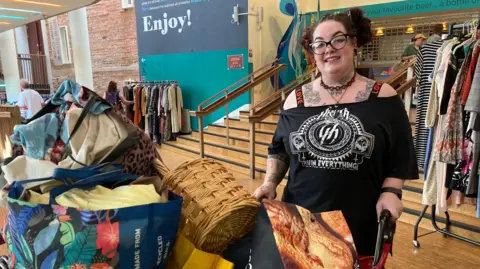 Alex Pope/BBC A woman, by a trolley piled high with items, wearing a black T-shirt, she is holding onto a mobility wheeled chair. She has tattoos and her hair is tied up. Clothes are behind her on rails. She is smiling and has glasses on and a necklace.