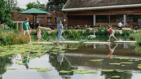 WWT An image of the trust showing a lake with wooden "lily pads" connecting both sides. There is a man and a woman and two children walking across the lily pads. On the lake there is also a swan. Behind the lake, there are multiple benches and umbrella, with the restaurant behind.