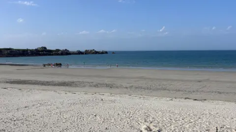 BBC The image is of the beach where you can see the blue sky with clouds, the sea water, some land and rocks on the left, and sand. 