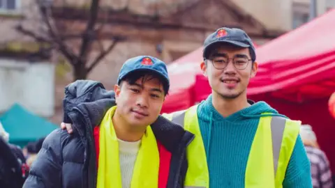 Josh (left) with black hair wearing a dark blue coat and white jumper with a lime green scarf and blue baseball cap with a Lancashire red rose logo stands in Lancaster city centre with Percy, who has his arm around Josh's shoulder. Percy is wearing a blue knitted hooded jumper and a yellow hi-vis vest. He has black hair and is wearing an identical blue baseball cap with a Lancashire red rose logo. You can see red stalls as part of the Chinese New Year festival. They are both smiling.
