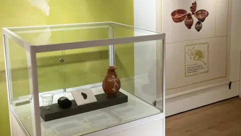 James Grant/BBC A glass cabinet containing a glass cup, two small urns and a shard of glass.