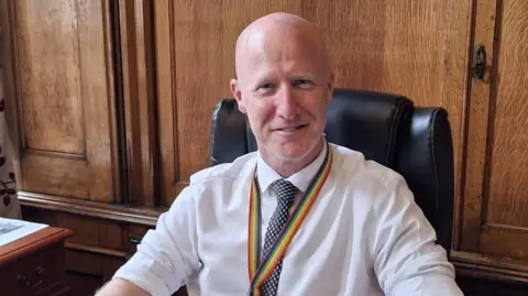 Lee Ratcliffe is wearing a white shirt with the sleeves rolled up and a blue and white patterned tie