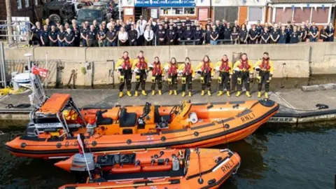 Pair of orange lifeboats side by side along harbour wall a line of RNLI crew stand along the side of the harbour wall.