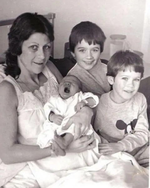 A woman in a hospital bed holdin a baby with two other young boys at the bedside. 